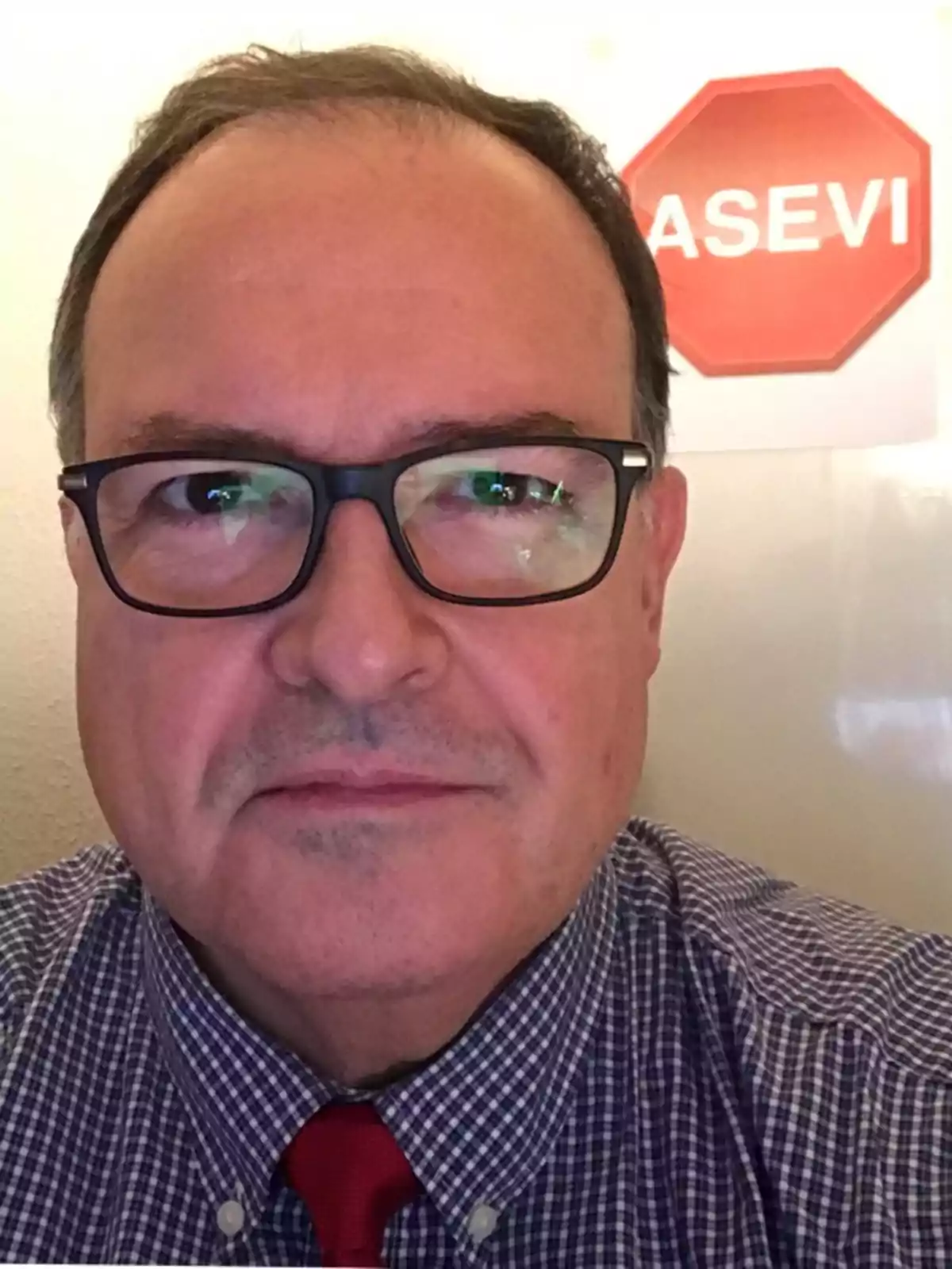 Hombre de mediana edad con gafas y camisa a cuadros, lleva una corbata roja y detrás de él hay un cartel con la palabra ASEVI sobre un fondo en forma de señal de alto Hombre de mediana edad con gafas y camisa a cuadros, lleva una corbata roja y detrás de él hay un cartel con la palabra ASEVI sobre un fondo en forma de señal de alto