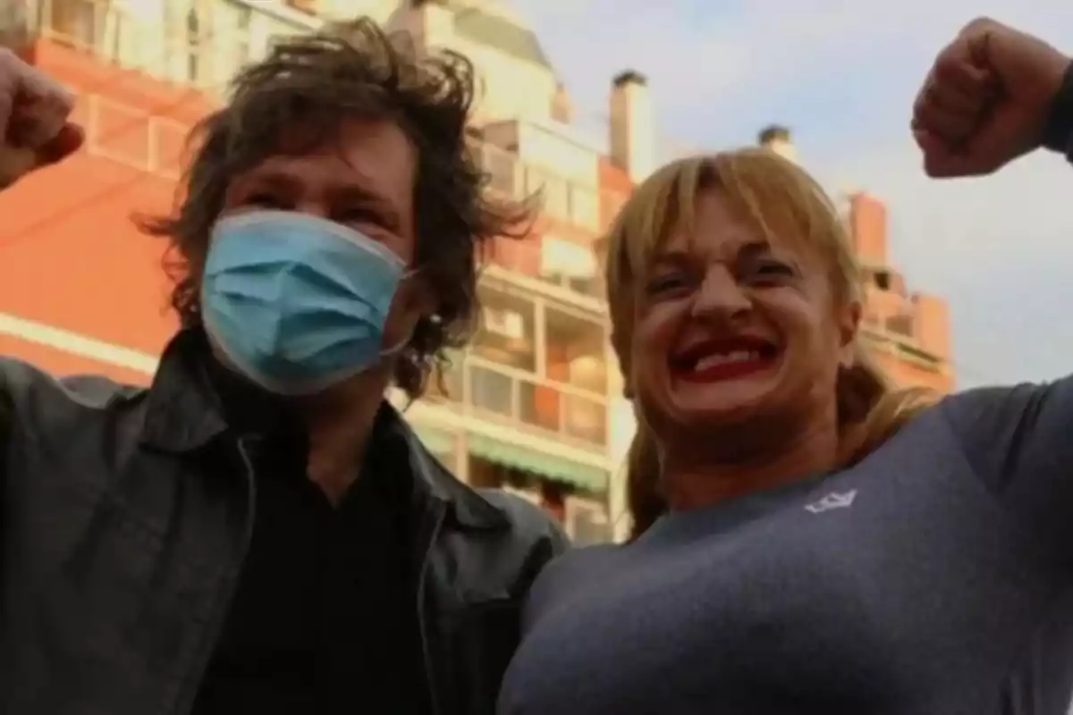 Dos personas levantan los brazos en señal de victoria una de ellas lleva mascarilla y la otra sonríe ampliamente con edificios de fondo