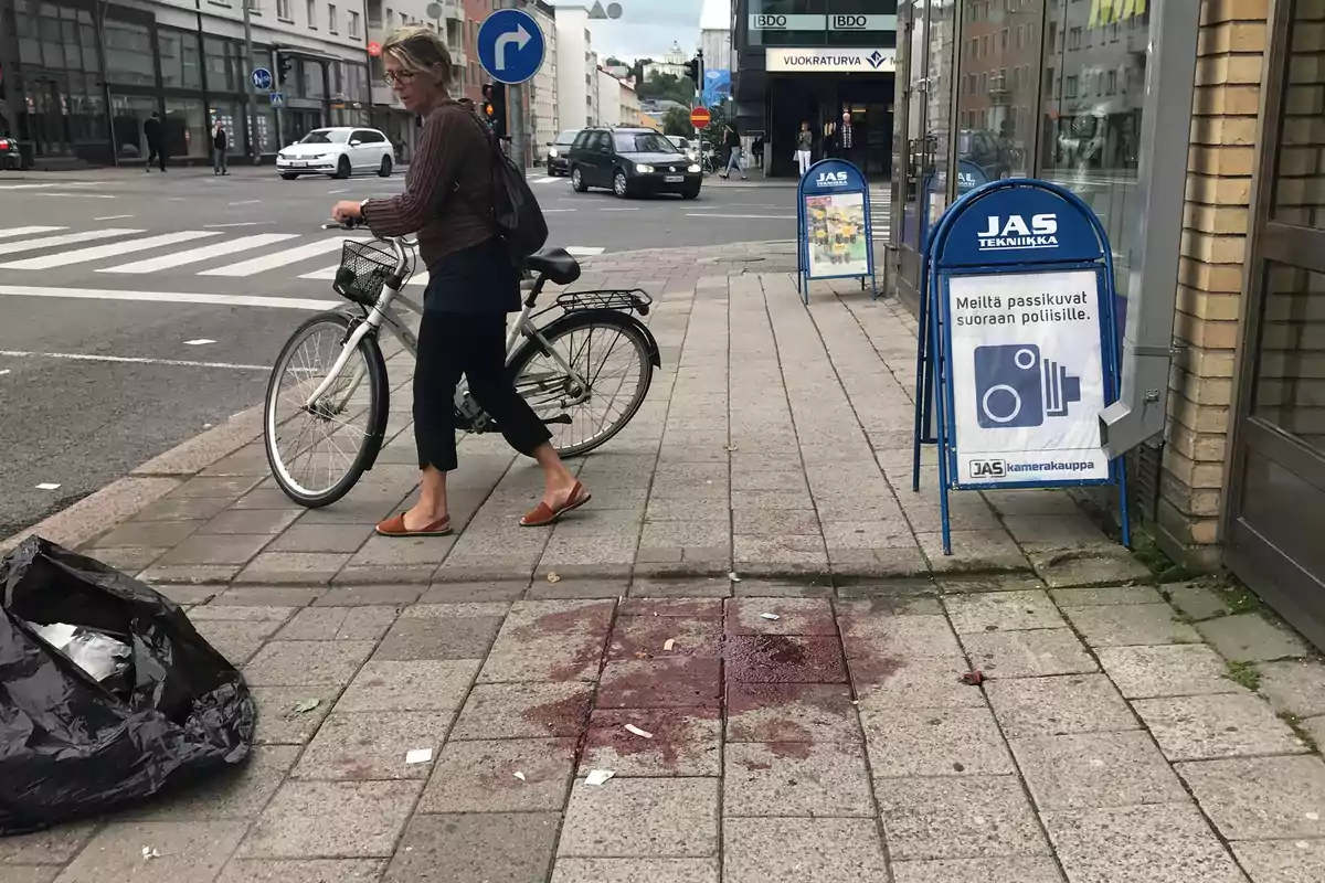 Una mujer camina empujando una bicicleta por la acera de una ciudad junto a una bolsa de basura negra y una mancha oscura en el suelo Una mujer camina empujando una bicicleta por la acera de una ciudad junto a una bolsa de basura negra y una mancha oscura en el suelo