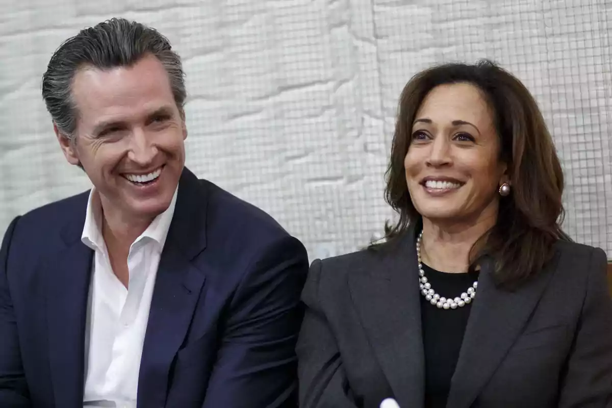 Two elegantly dressed people smile while sitting next to each other against a light background.
