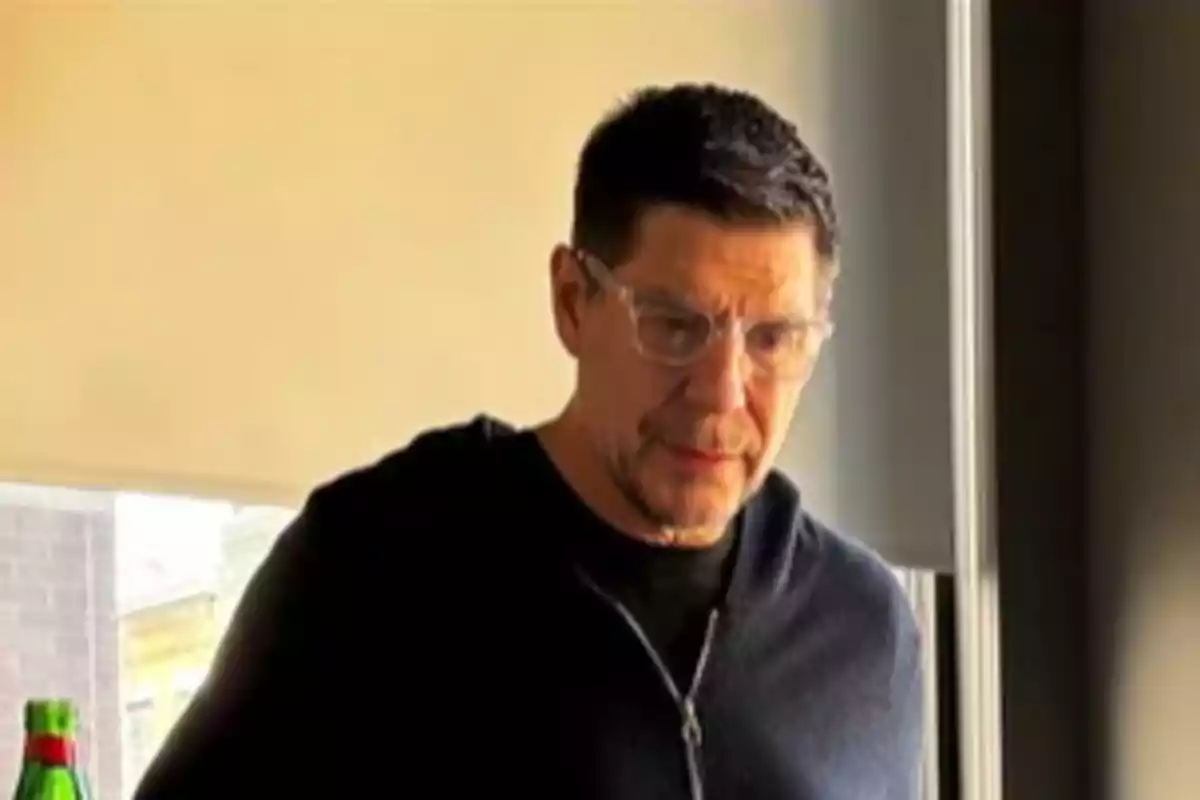 Hombre de cabello corto y gafas transparentes con suéter oscuro de pie junto a una ventana iluminada por luz natural Hombre de cabello corto y gafas transparentes con suéter oscuro de pie junto a una ventana iluminada por luz natural