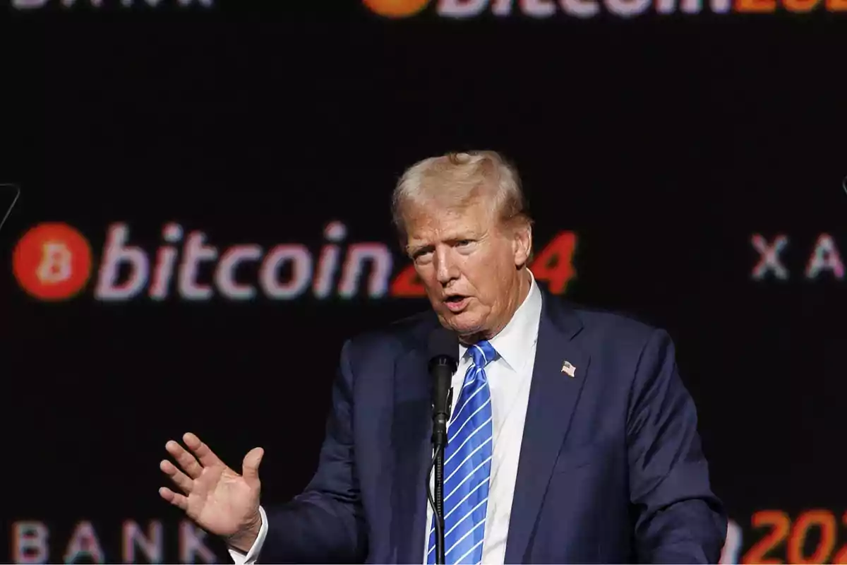 Hombre de cabello rubio con traje azul y corbata azul hablando frente a un micrófono con el logo de bitcoin y el número 2024 en el fondo