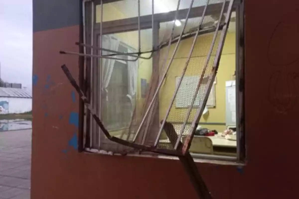 Ventana de un edificio con la reja doblada y dañada mostrando el interior de un aula iluminada Ventana de un edificio con la reja doblada y dañada mostrando el interior de un aula iluminada