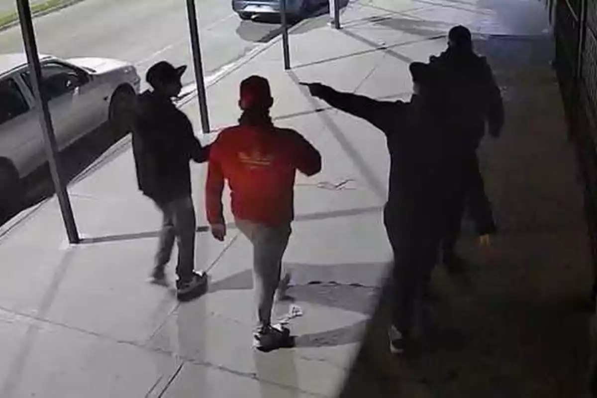 Cuatro personas caminando por una acera de noche, una de ellas apunta con un objeto a las otras tres Cuatro personas caminando por una acera de noche, una de ellas apunta con un objeto a las otras tres
