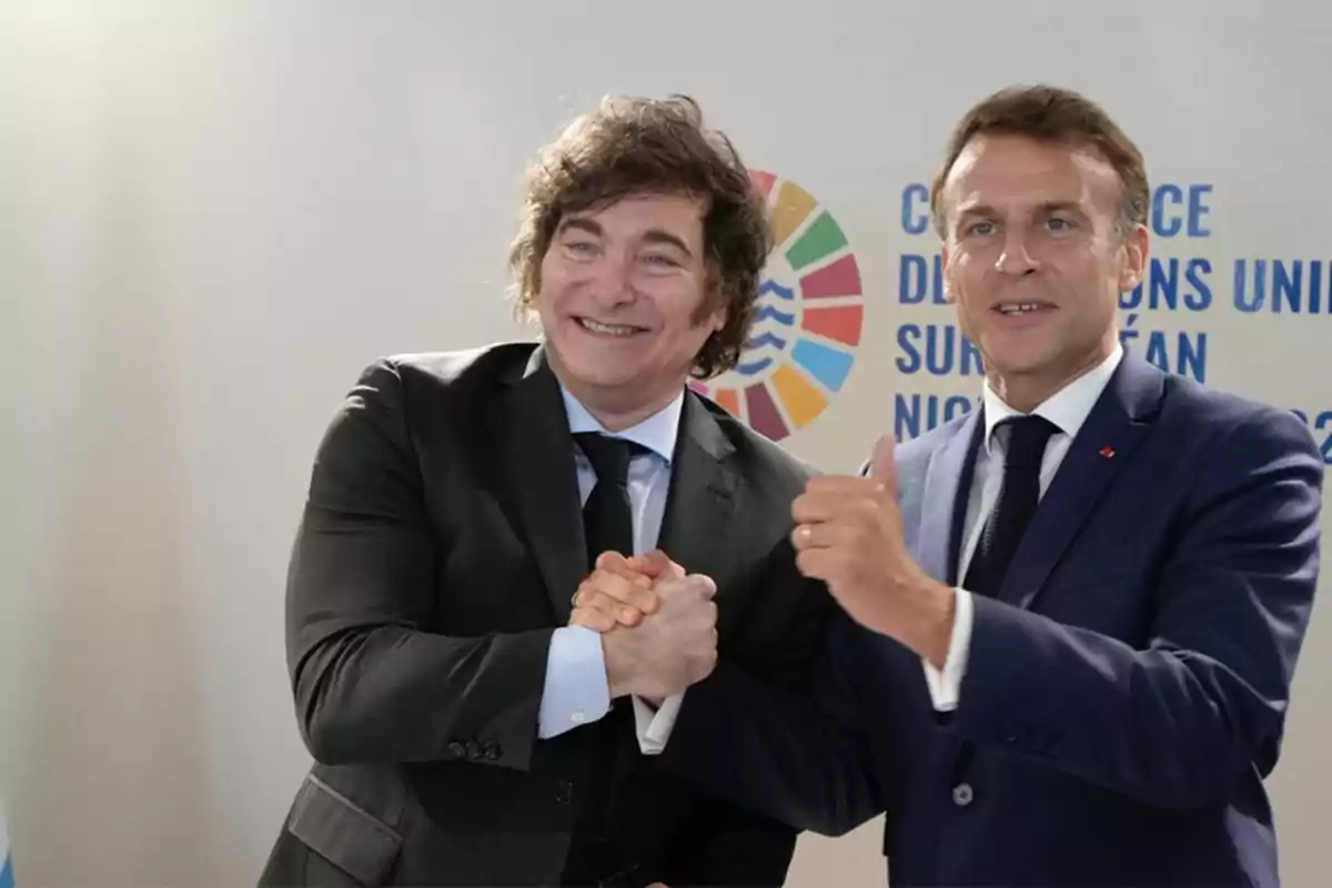 Dos hombres vestidos con traje oscuro se saludan sonrientes y posan para la cámara frente a un fondo con logotipos y texto institucional. Dos hombres vestidos con traje oscuro se saludan sonrientes y posan para la cámara frente a un fondo con logotipos y texto institucional.