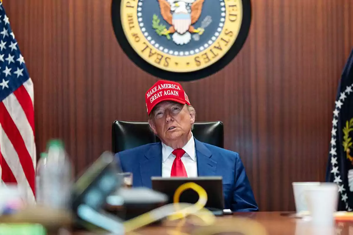 Hombre con traje azul y corbata roja sentado en una mesa con una gorra roja que dice Make America Great Again y el escudo presidencial de Estados Unidos en la pared detrás de él