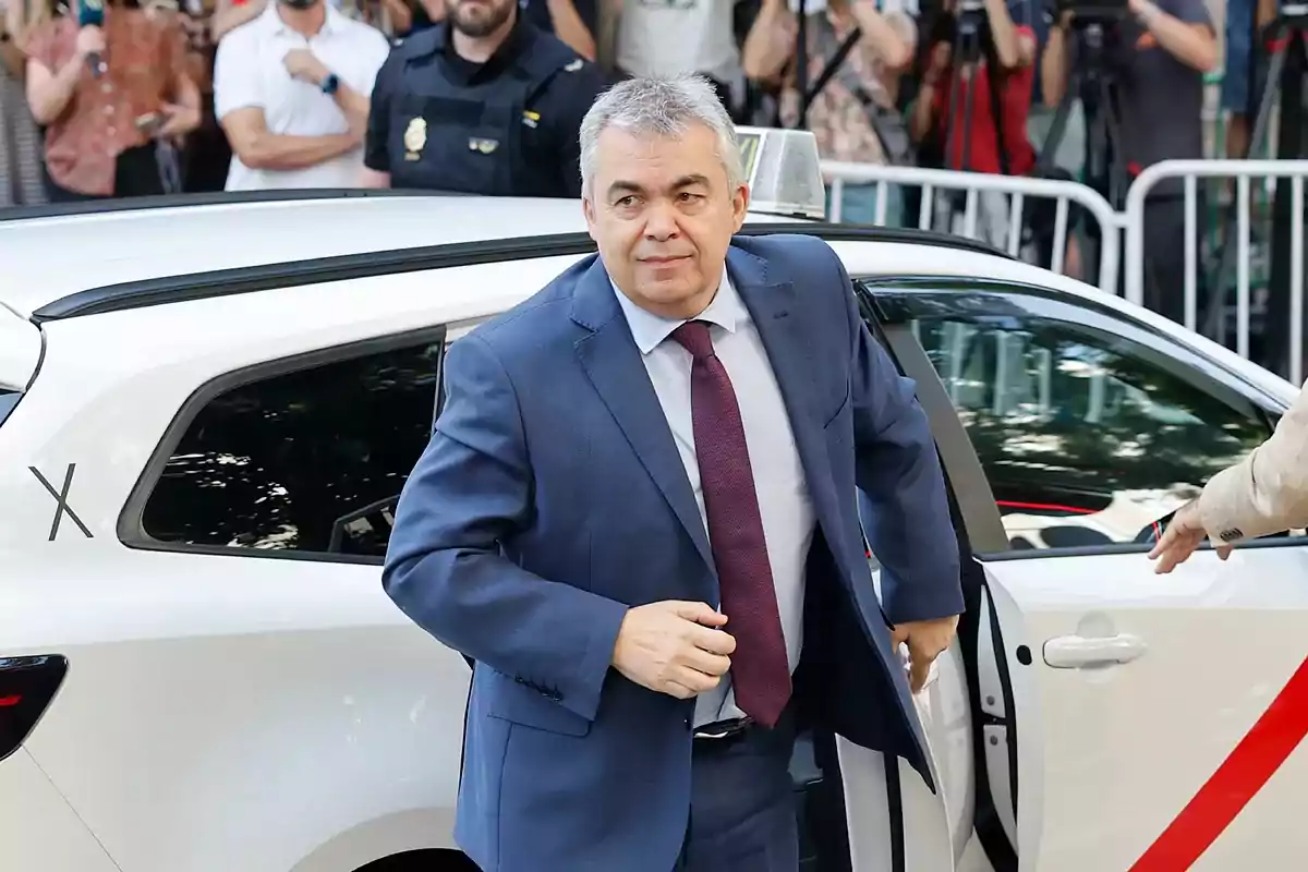 Hombre de cabello canoso con traje azul y corbata roja bajando de un taxi blanco mientras varias personas y policías observan al fondo