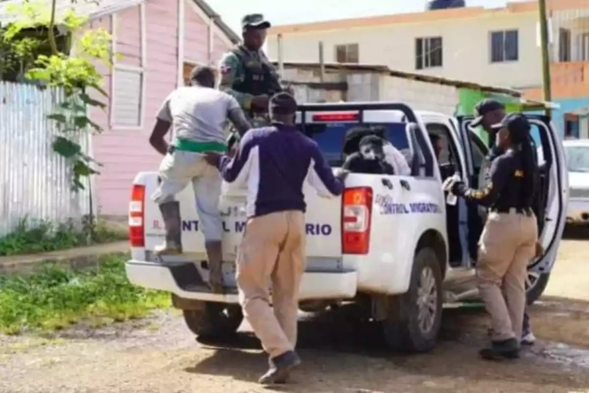 Varias personas están siendo escoltadas por agentes hacia la parte trasera de una camioneta blanca de control migratorio en una zona residencial.