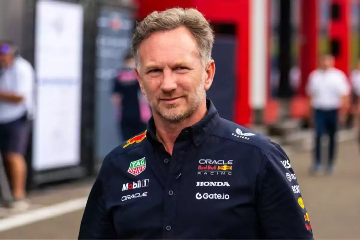 Hombre de mediana edad con barba y cabello corto, vistiendo una camisa azul marino con logotipos de patrocinadores de un equipo de automovilismo, de pie en un entorno al aire libre con personas y estructuras desenfocadas al fondo.