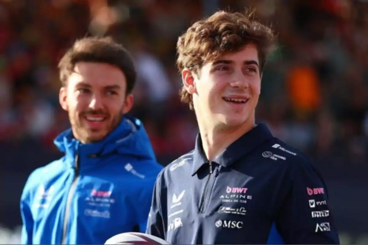 Two young people in motorsport team sportswear smile outdoors during the day.