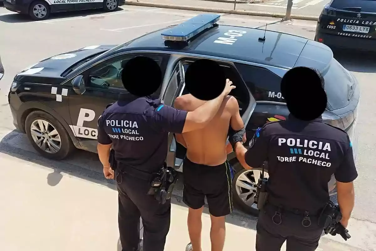 Dos policías locales escoltan a un hombre sin camiseta hacia un coche patrulla en una calle durante el día Dos policías locales escoltan a un hombre sin camiseta hacia un coche patrulla en una calle durante el día