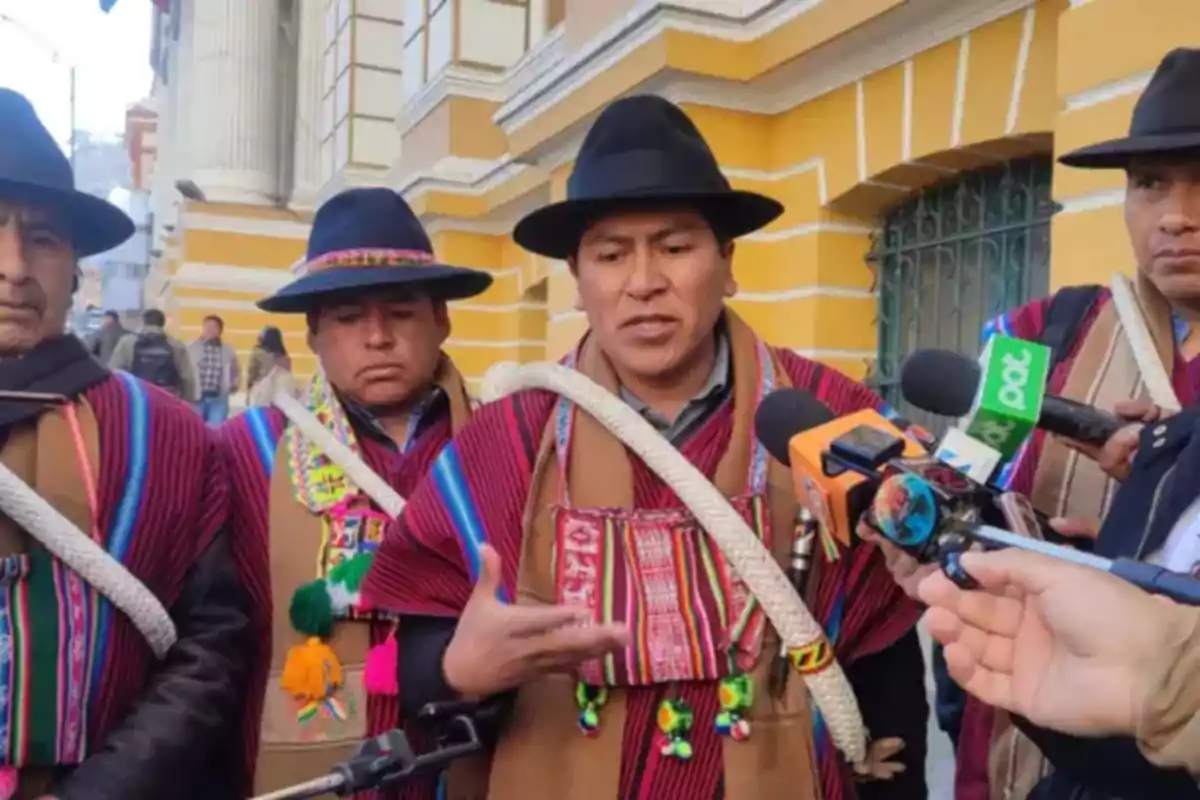 Cuatro hombres con vestimenta tradicional andina y sombreros negros dan declaraciones a la prensa frente a un edificio amarillo mientras varios micrófonos los rodean.
