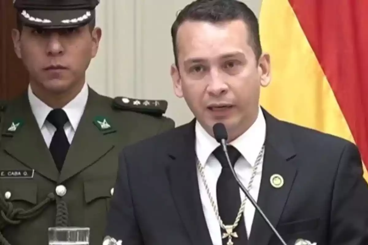 A man in a dark suit and tie speaks into a microphone while a uniformed military officer stands beside him, and a red and yellow flag can be seen in the background. A man in a dark suit and tie speaks into a microphone while a uniformed military officer stands beside him, and a red and yellow flag can be seen in the background.