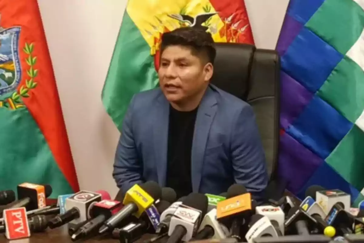 Man in a blue suit speaking at a press conference in front of several microphones and Bolivian flags in the background