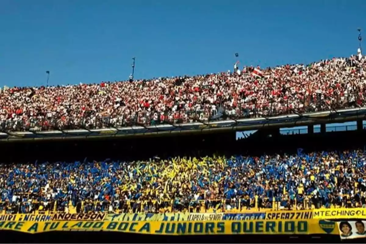 Aficionados de dos equipos de fútbol llenan las gradas de un estadio, unos vestidos de blanco y rojo y otros de azul y amarillo, separados por una reja y una bandera amarilla con letras azules.