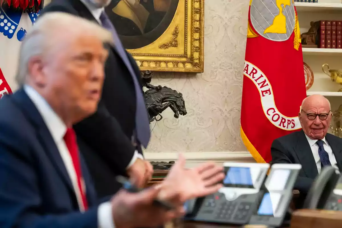 A gray-haired man with glasses sits next to a Marine Corps flag in an elegant office while another blurred person gestures in the foreground. A gray-haired man with glasses sits next to a Marine Corps flag in an elegant office while another blurred person gestures in the foreground.