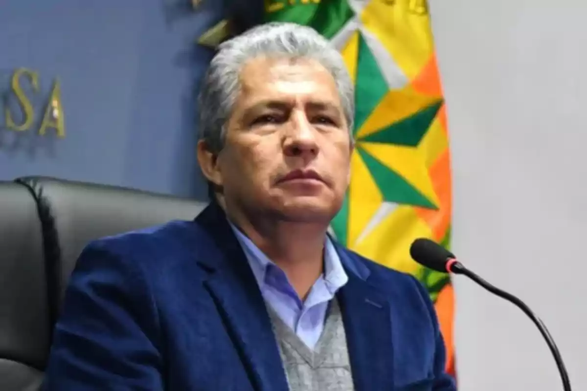 Hombre de cabello canoso con saco azul sentado frente a un micrófono y una bandera de fondo Hombre de cabello canoso con saco azul sentado frente a un micrófono y una bandera de fondo