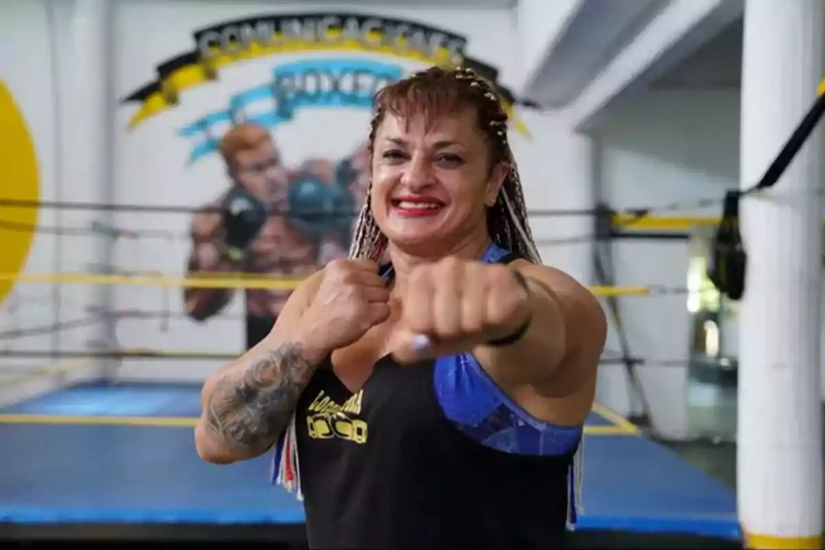 Mujer sonriente con trenzas y tatuaje en el brazo posa con el puño extendido en un gimnasio de boxeo Mujer sonriente con trenzas y tatuaje en el brazo posa con el puño extendido en un gimnasio de boxeo