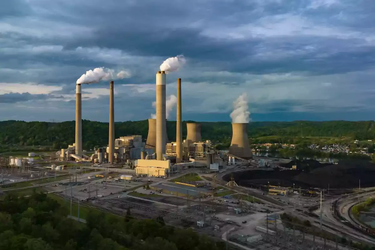 Vista aérea de una planta industrial con varias chimeneas expulsando humo y torres de enfriamiento rodeada de vegetación y cielo nublado Vista aérea de una planta industrial con varias chimeneas expulsando humo y torres de enfriamiento rodeada de vegetación y cielo nublado