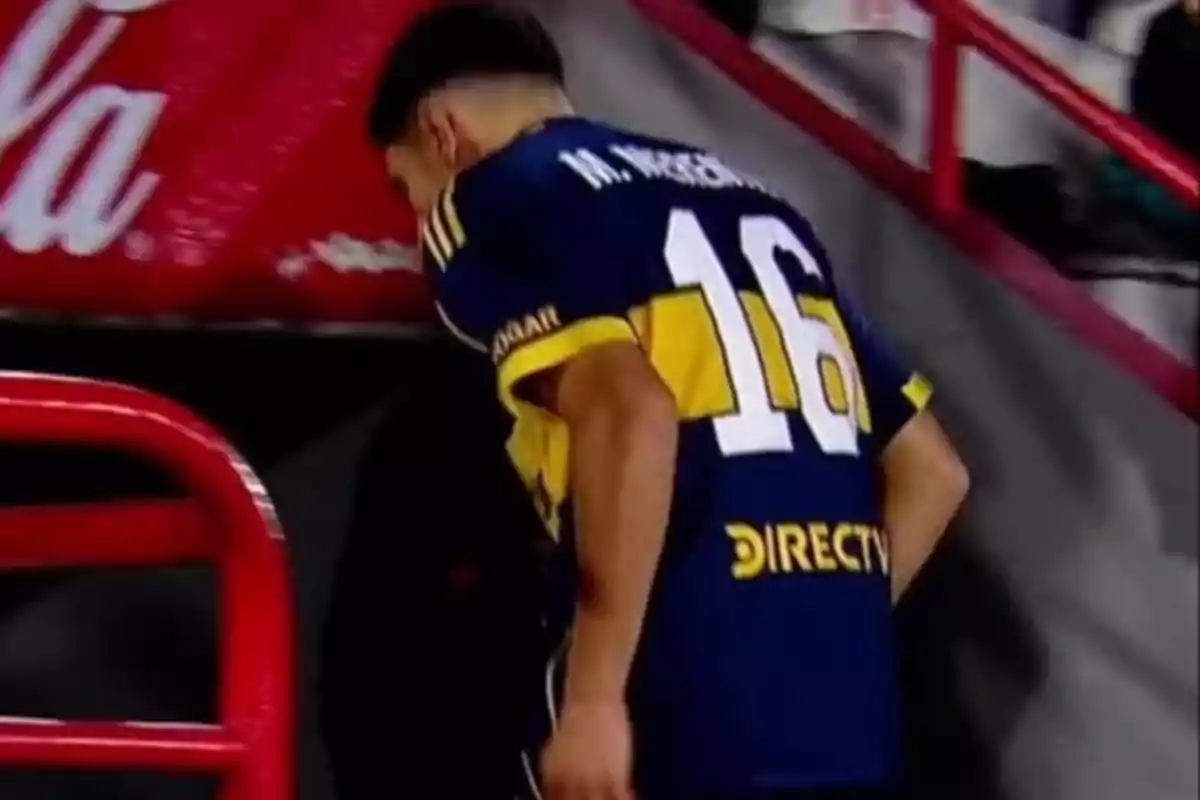 Jugador de fútbol con camiseta azul y amarilla número 16 caminando cabizbajo cerca de una baranda roja en un estadio Jugador de fútbol con camiseta azul y amarilla número 16 caminando cabizbajo cerca de una baranda roja en un estadio
