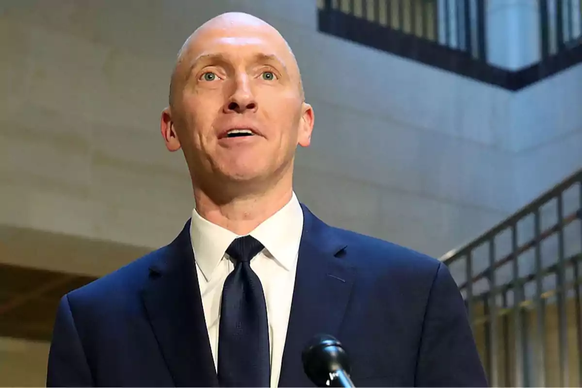 Bald man in a dark blue suit and tie speaking in front of a microphone in a building with stairs and railings in the background Bald man in a dark blue suit and tie speaking in front of a microphone in a building with stairs and railings in the background