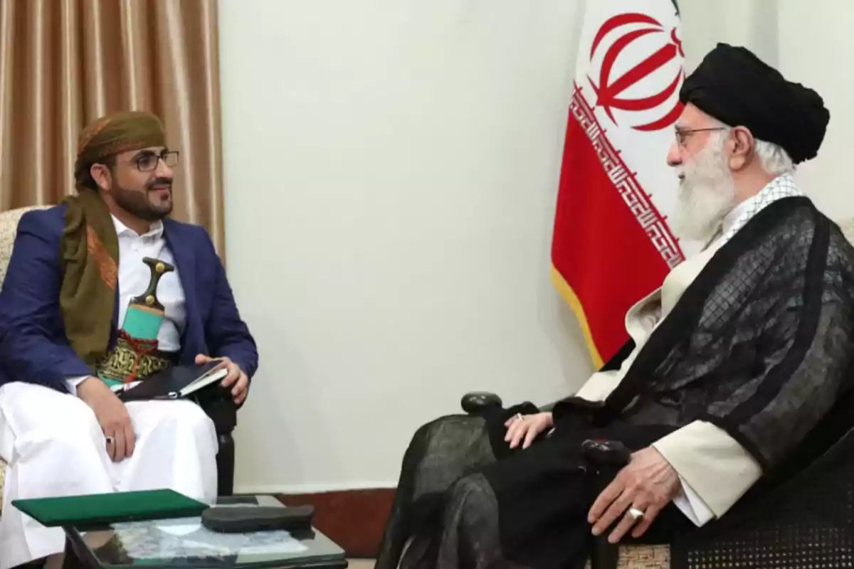 Dos hombres sentados conversan en una sala, uno lleva vestimenta tradicional yemení y el otro viste ropa tradicional iraní, con una bandera de Irán al fondo.