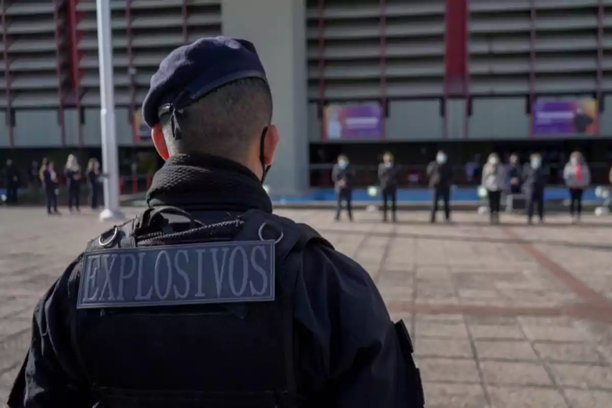 A bomb squad agent with his back turned watches a group of people in front of a building. A bomb squad agent with his back turned watches a group of people in front of a building.