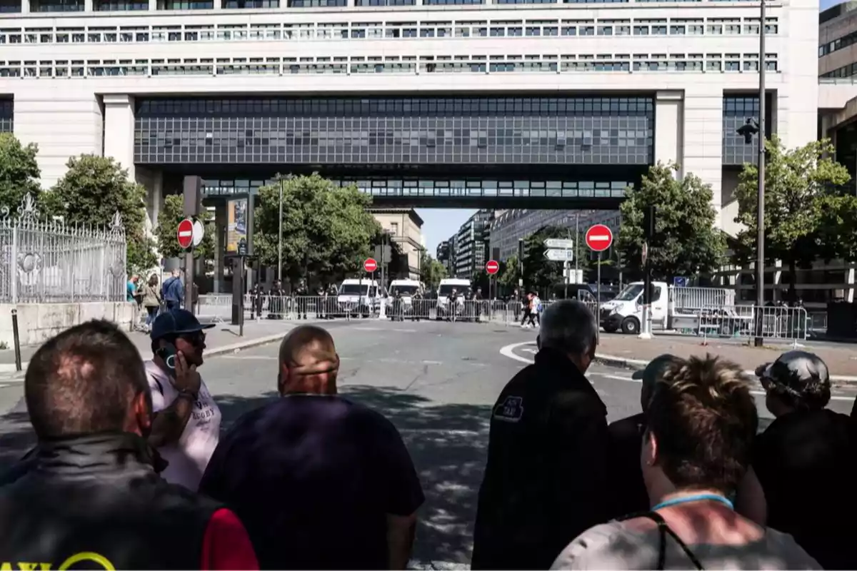 Varias personas observan una calle bloqueada por vallas y vehículos policiales frente a un edificio moderno con pasarelas elevadas y árboles a los lados. Varias personas observan una calle bloqueada por vallas y vehículos policiales frente a un edificio moderno con pasarelas elevadas y árboles a los lados.