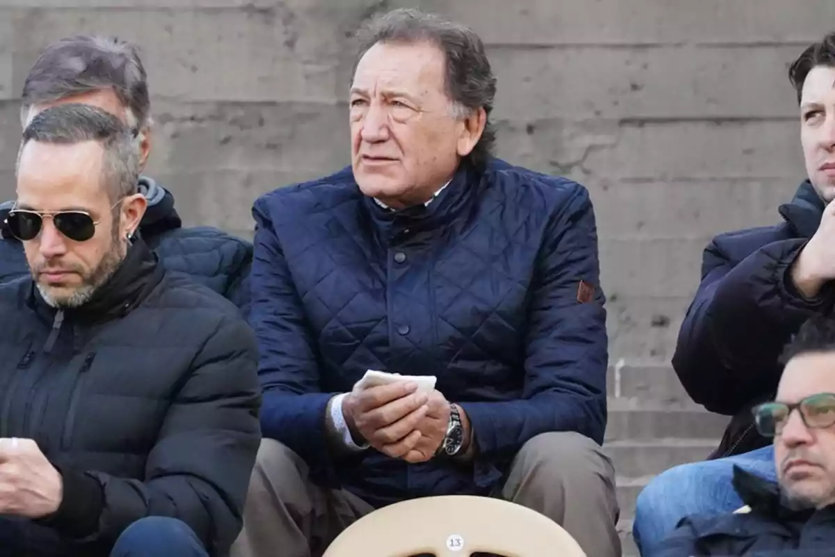 Un hombre de cabello canoso y chaqueta azul acolchada está sentado en unas gradas junto a otras personas, observando algo al frente con expresión seria.