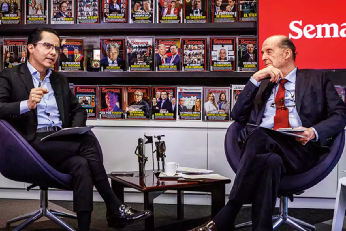 Dos hombres de traje conversan sentados en sillas modernas frente a una mesa pequeña en una oficina decorada con portadas de la revista Semana