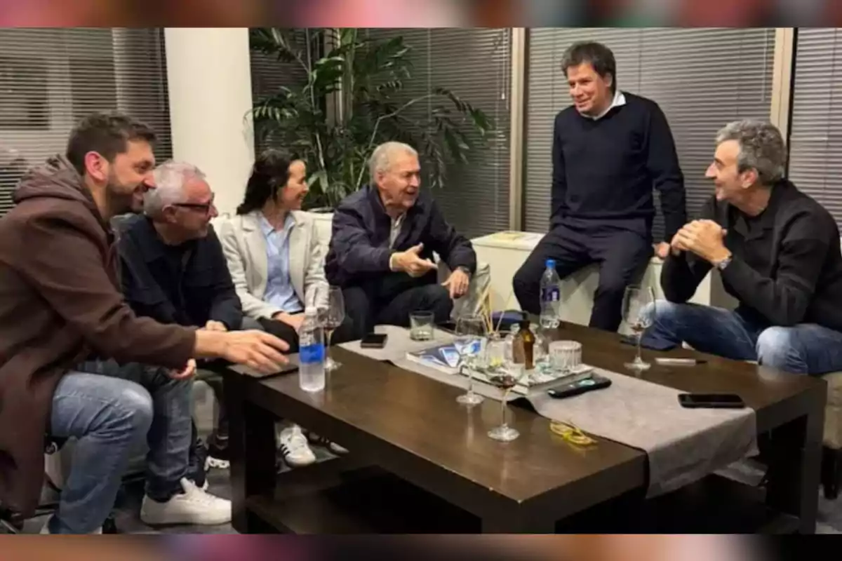 Seis personas conversan y ríen sentadas alrededor de una mesa de madera con copas, botellas y objetos personales en una sala iluminada con plantas al fondo. Seis personas conversan y ríen sentadas alrededor de una mesa de madera con copas, botellas y objetos personales en una sala iluminada con plantas al fondo.