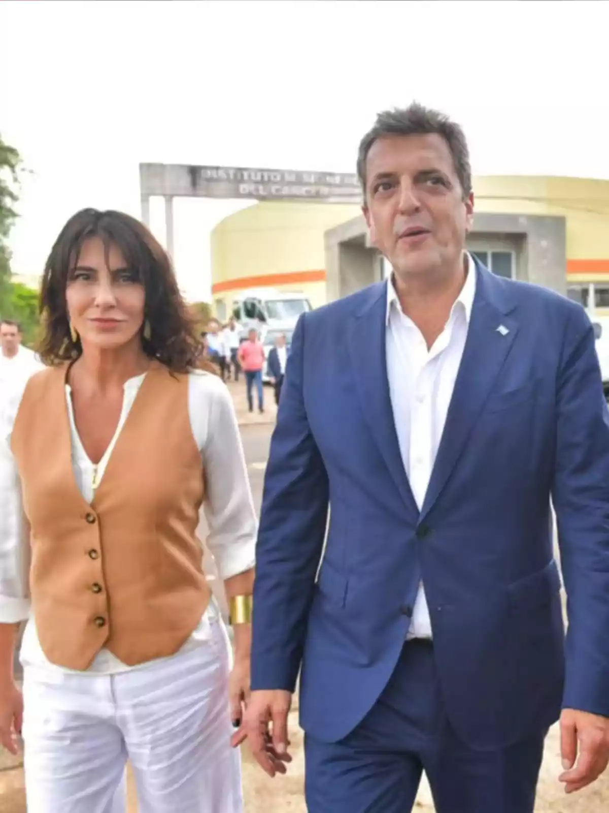 Una mujer con chaleco marrón y pantalón blanco camina junto a un hombre con traje azul frente a un edificio con un cartel que dice Instituto Misionero del Cáncer Una mujer con chaleco marrón y pantalón blanco camina junto a un hombre con traje azul frente a un edificio con un cartel que dice Instituto Misionero del Cáncer