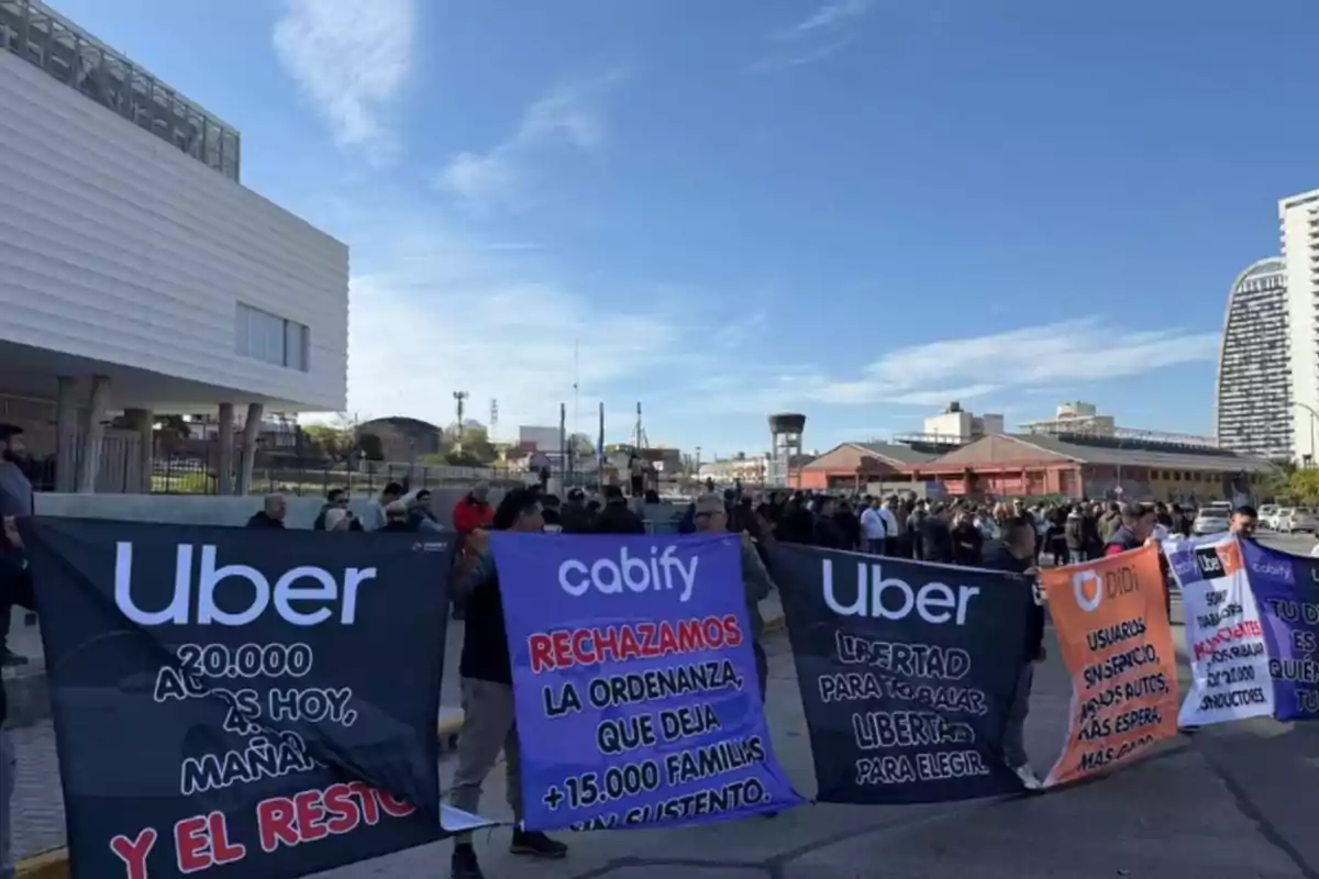Personas manifestándose en la calle con pancartas de Uber, Cabify y Didi frente a un edificio moderno y bajo un cielo despejado Personas manifestándose en la calle con pancartas de Uber, Cabify y Didi frente a un edificio moderno y bajo un cielo despejado
