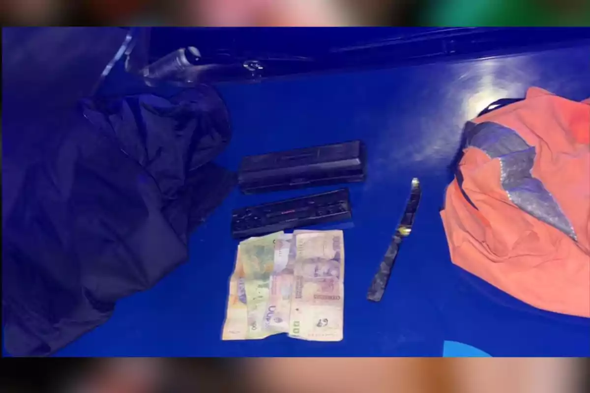 Sobre una superficie azul hay dos prendas de ropa, billetes, una navaja y dos estuches negros rectangulares. Sobre una superficie azul hay dos prendas de ropa, billetes, una navaja y dos estuches negros rectangulares.