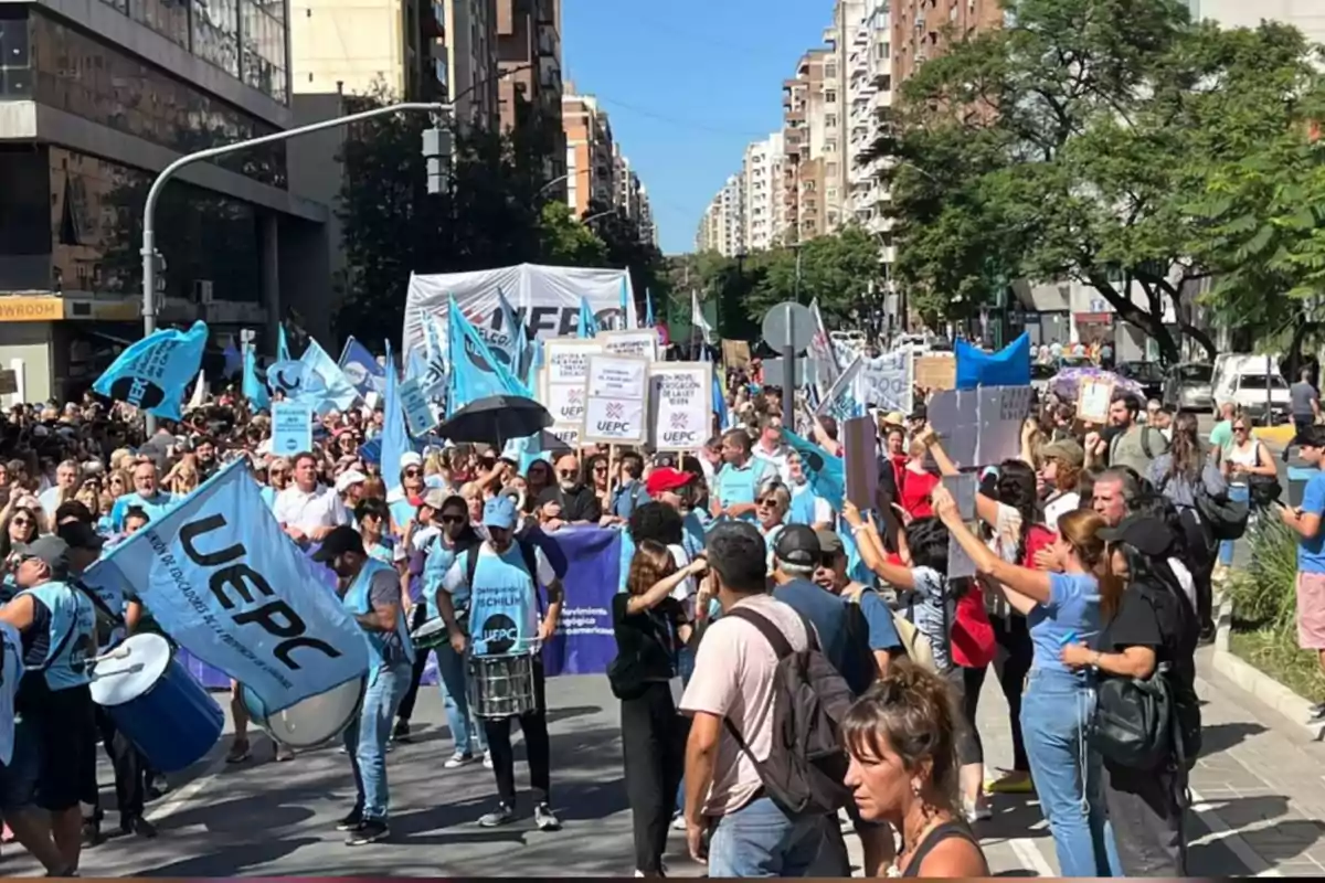 Manifestación multitudinaria en una avenida de la ciudad con personas portando banderas celestes, carteles y tambores, rodeadas de edificios y árboles Manifestación multitudinaria en una avenida de la ciudad con personas portando banderas celestes, carteles y tambores, rodeadas de edificios y árboles