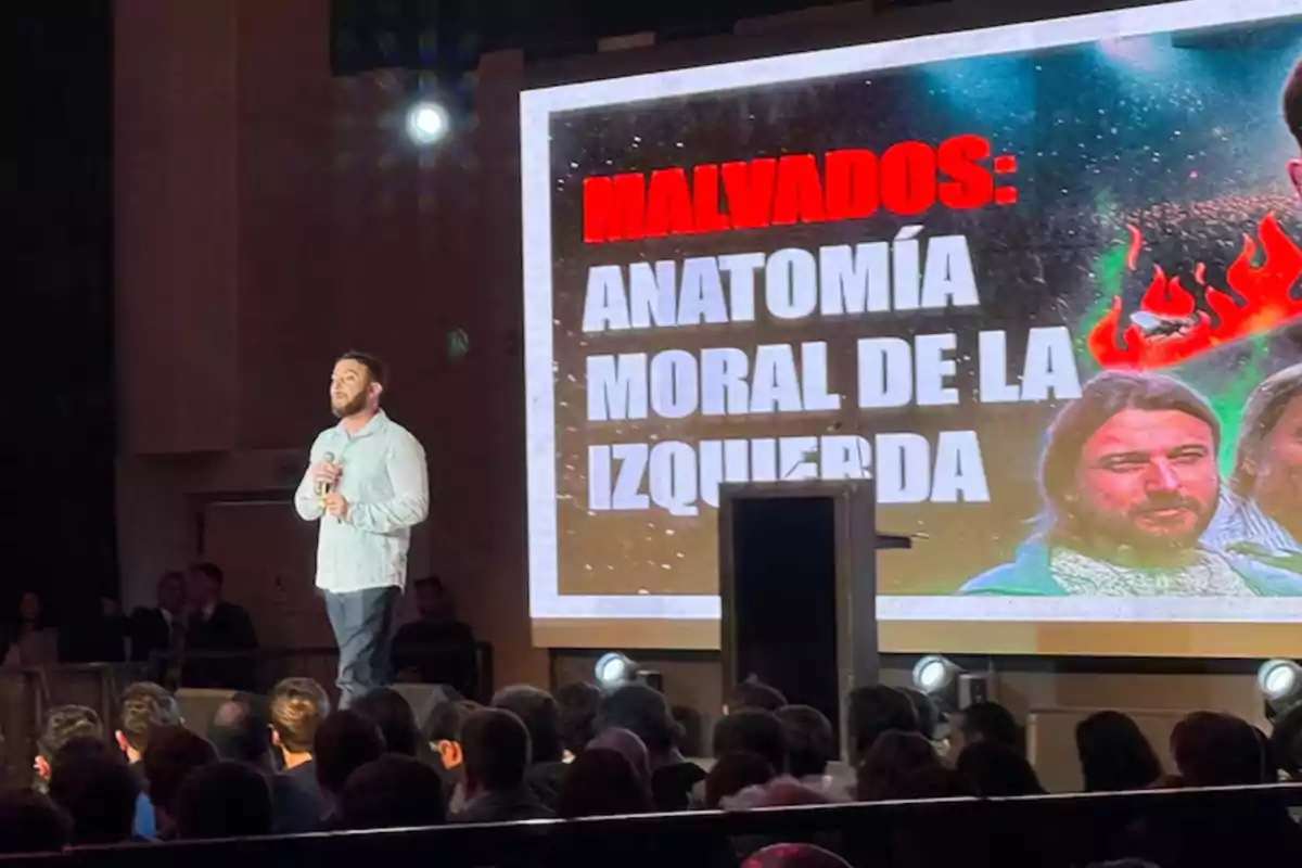 Un hombre de pie con micrófono frente a una audiencia, junto a una pantalla grande que muestra el texto Malvados Anatomía moral de la izquierda y la imagen de dos personas