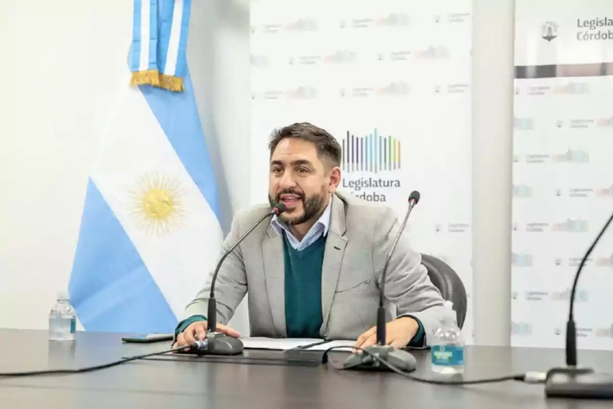 Bearded man in a gray jacket speaking at a press conference in the Córdoba Legislature with an Argentine flag in the background Bearded man in a gray jacket speaking at a press conference in the Córdoba Legislature with an Argentine flag in the background