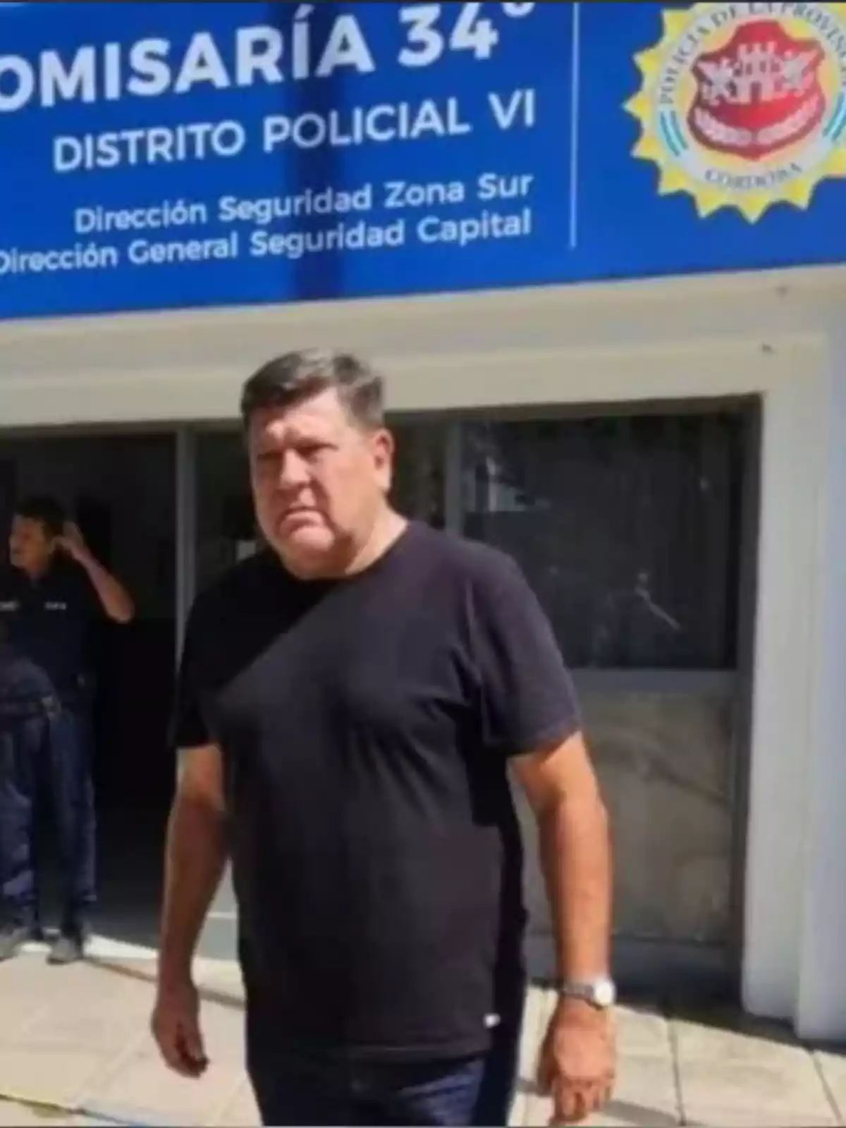Hombre de camiseta negra de pie frente a una comisaría con un cartel azul en la entrada Hombre de camiseta negra de pie frente a una comisaría con un cartel azul en la entrada