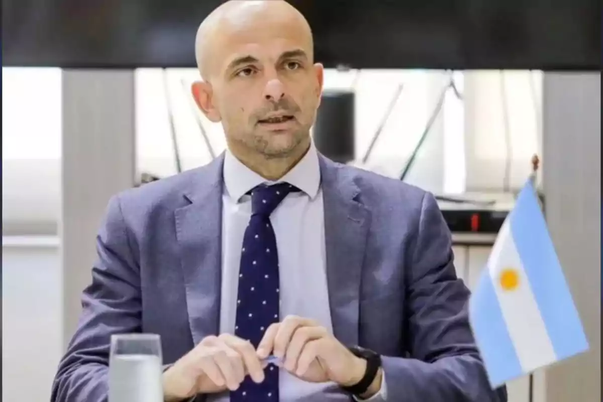 Hombre calvo con traje y corbata azul sentado en una mesa junto a una bandera de Argentina y un vaso de agua Hombre calvo con traje y corbata azul sentado en una mesa junto a una bandera de Argentina y un vaso de agua