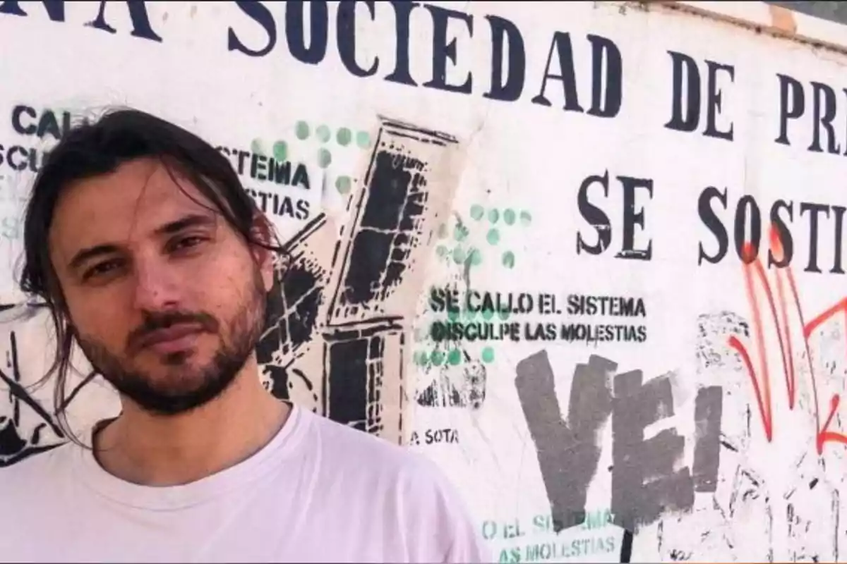 Hombre de cabello largo y barba con camiseta blanca posando frente a una pared con grafitis y un cartel con letras grandes y negras Hombre de cabello largo y barba con camiseta blanca posando frente a una pared con grafitis y un cartel con letras grandes y negras