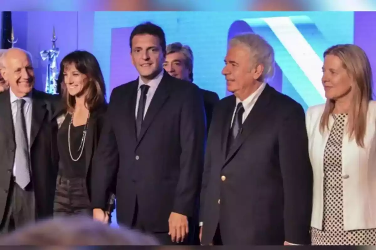 Cinco personas vestidas de manera formal posan juntas y sonríen en un evento con fondo azul. Cinco personas vestidas de manera formal posan juntas y sonríen en un evento con fondo azul.
