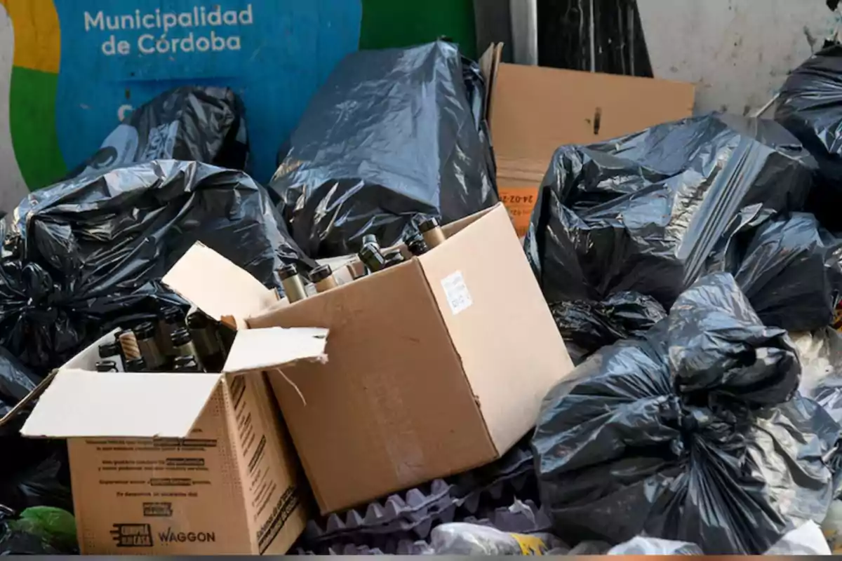 Cajas de cartón llenas de botellas de vidrio y bolsas de basura negras apiladas en la calle frente a un contenedor de reciclaje de la Municipalidad de Córdoba Cajas de cartón llenas de botellas de vidrio y bolsas de basura negras apiladas en la calle frente a un contenedor de reciclaje de la Municipalidad de Córdoba