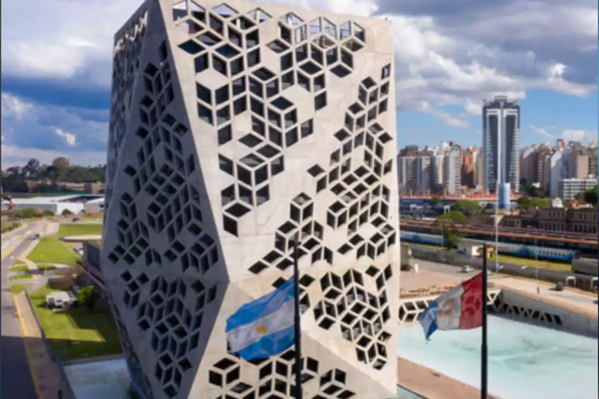 Edificio moderno con diseño geométrico en la fachada, banderas ondeando al frente y una ciudad al fondo bajo un cielo parcialmente nublado