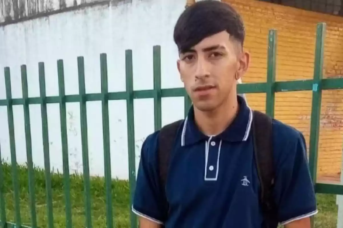 Joven con cabello corto y oscuro viste una camiseta tipo polo azul marino y lleva una mochila negra de pie frente a una reja verde y una pared blanca y amarilla Joven con cabello corto y oscuro viste una camiseta tipo polo azul marino y lleva una mochila negra de pie frente a una reja verde y una pared blanca y amarilla