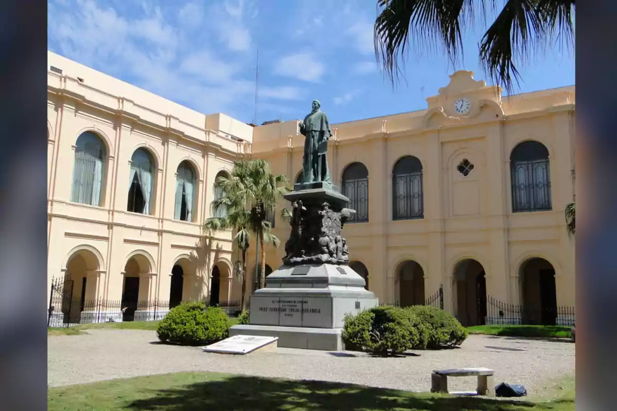 Estatua de un hombre sobre un pedestal en el centro de un patio rodeado por un edificio de fachada amarilla con ventanas arqueadas y un reloj en la parte superior Estatua de un hombre sobre un pedestal en el centro de un patio rodeado por un edificio de fachada amarilla con ventanas arqueadas y un reloj en la parte superior