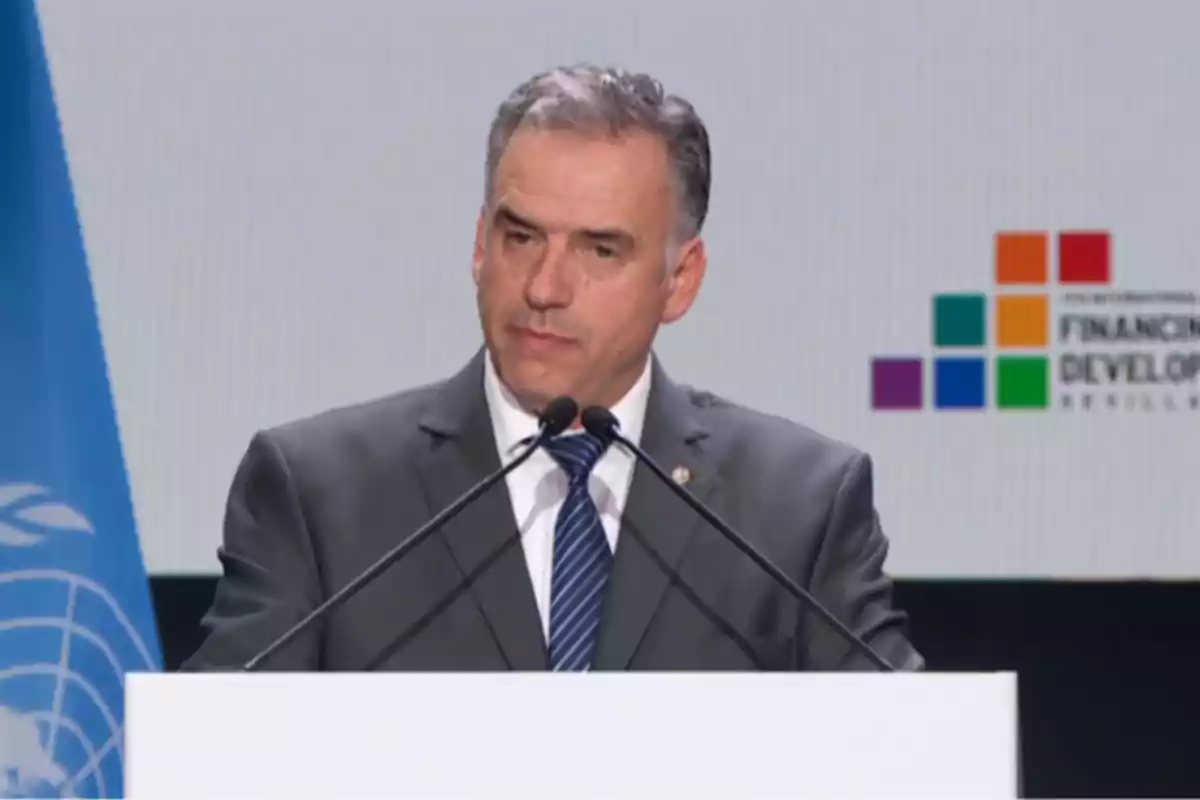 Man in a suit speaking at a podium with two microphones during an international event with a blue flag and a colorful logo in the background