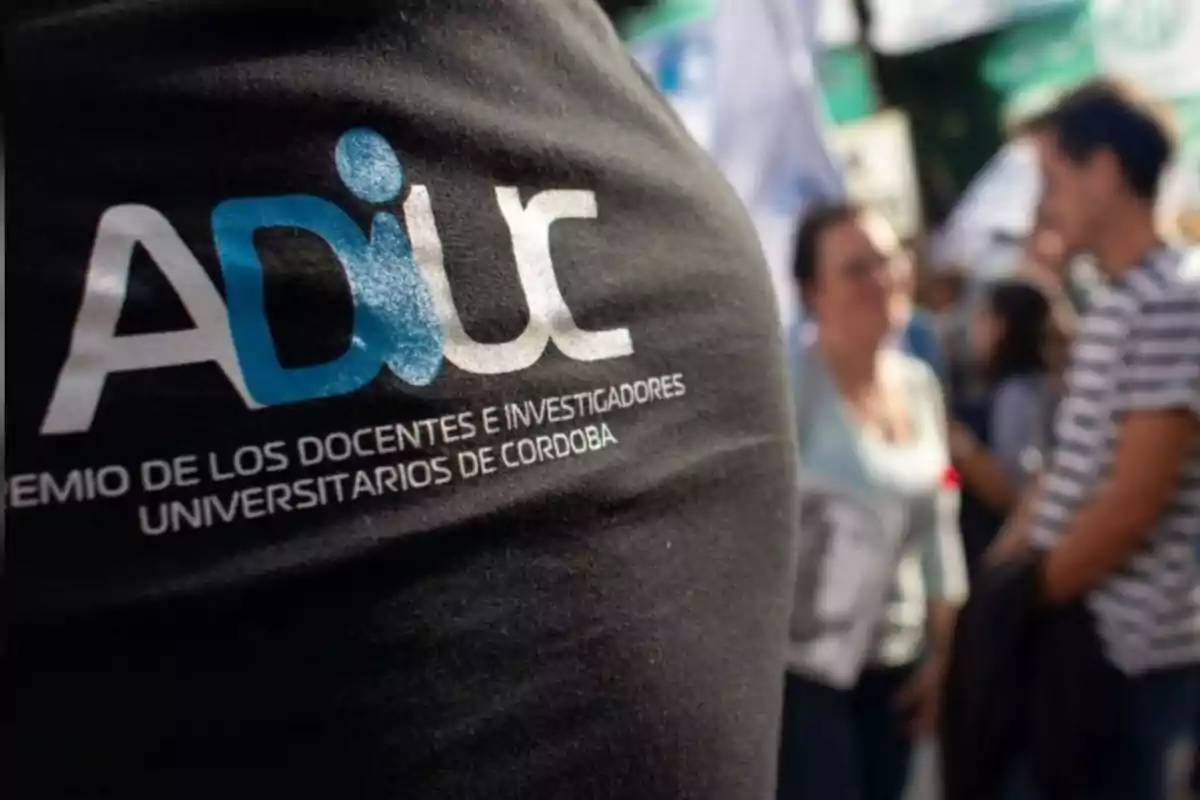 Persona con camiseta negra de ADIUC en una manifestación con gente y banderas al fondo Persona con camiseta negra de ADIUC en una manifestación con gente y banderas al fondo