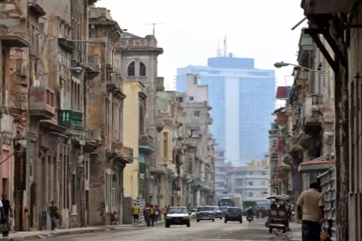 Calle urbana con edificios antiguos y desgastados a los lados, personas caminando y autos circulando, al fondo se observa un edificio moderno y alto