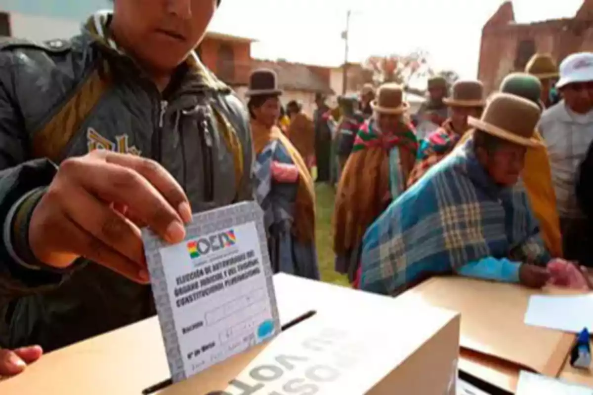 Persona depositando su voto en una urna mientras otras personas esperan en fila en un ambiente al aire libre