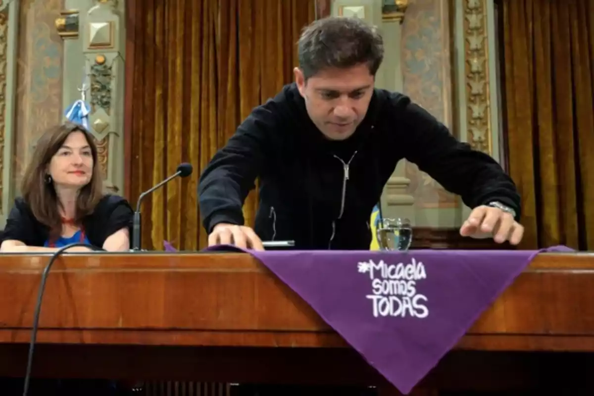 A man and a woman are sitting at a wooden table in a decorated room; on the table, there is a purple scarf with the phrase "Micaela somos todas."